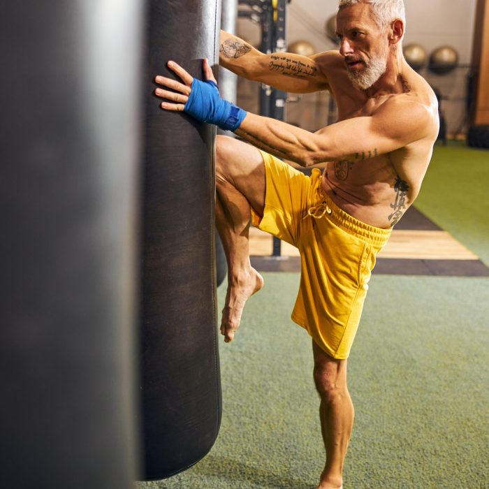 Full-sized portrait of a gray-headed bearded barefoot shirtless Caucasian male athlete kneeing the punching bag
